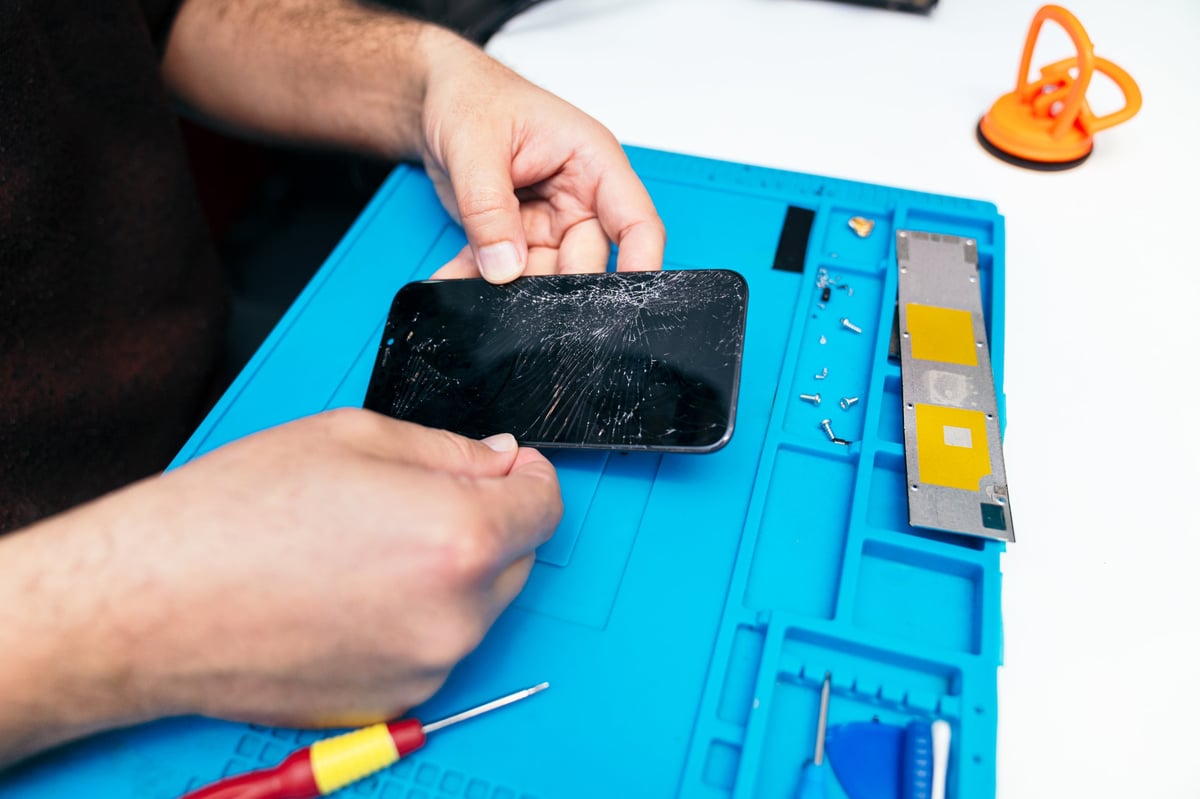 Händer som reparerar en iPhone med sprucken skärm på en blå matta